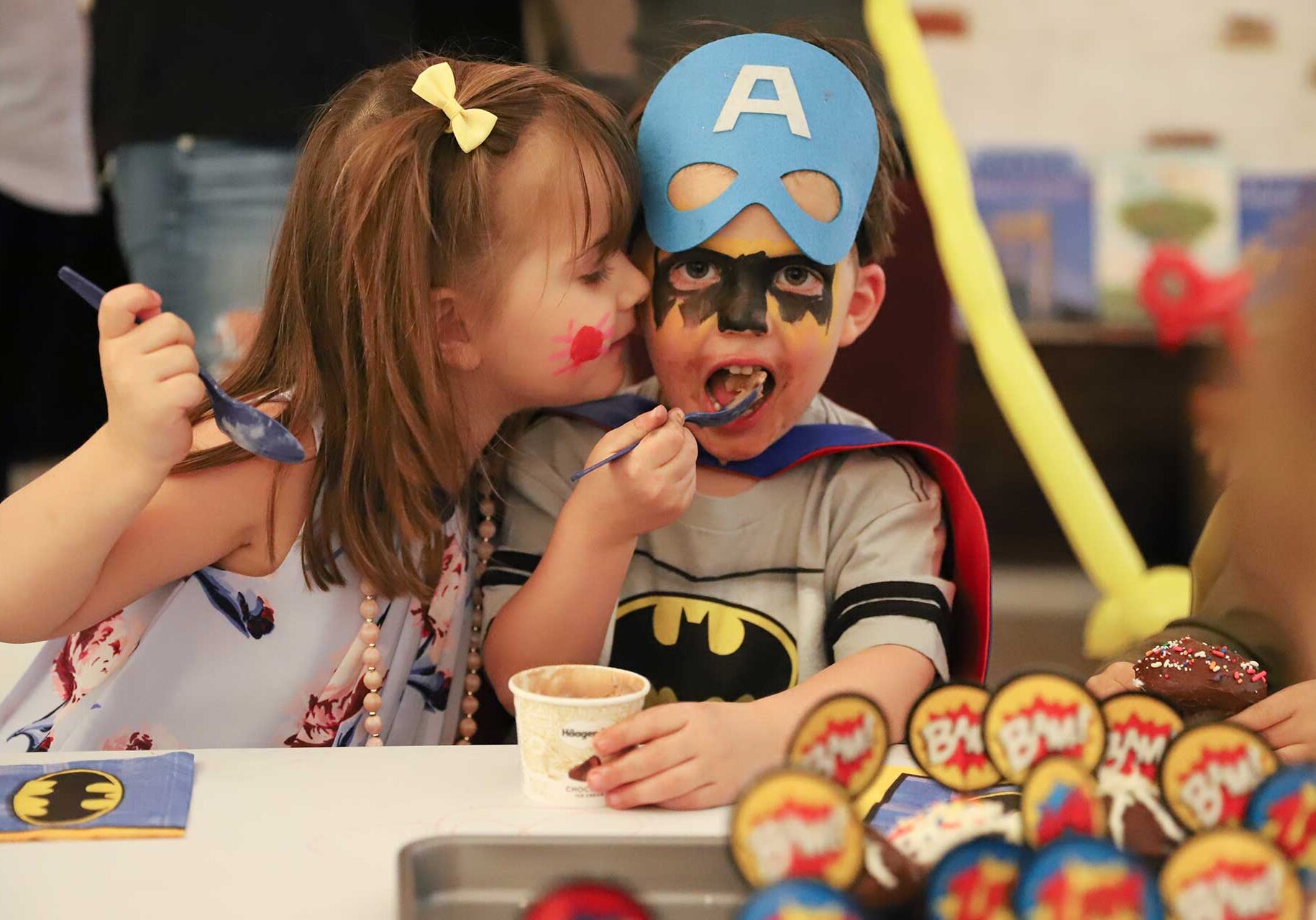 A boy and a girl wearing clown makeup and party clothes eating a dessert while the girl gives the boy a kiss on the cheek